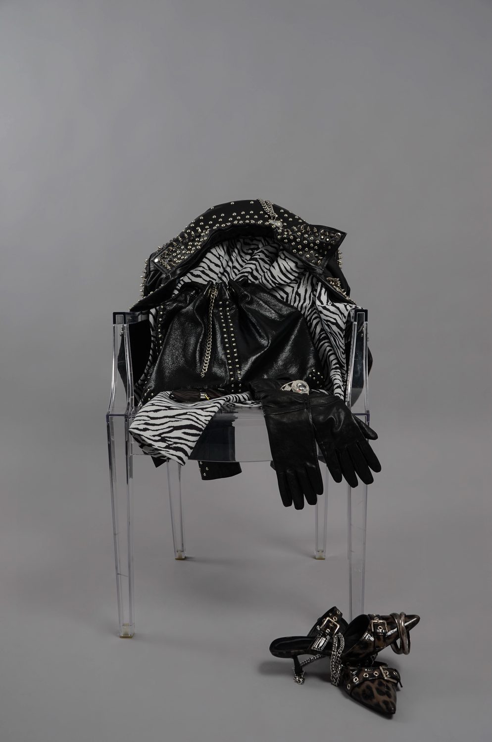 Chaos — a studded black leather jacket, zebra-print fabric and leather gloves heaped over a clear acrylic chair on a grey ground, printed heels fallen below