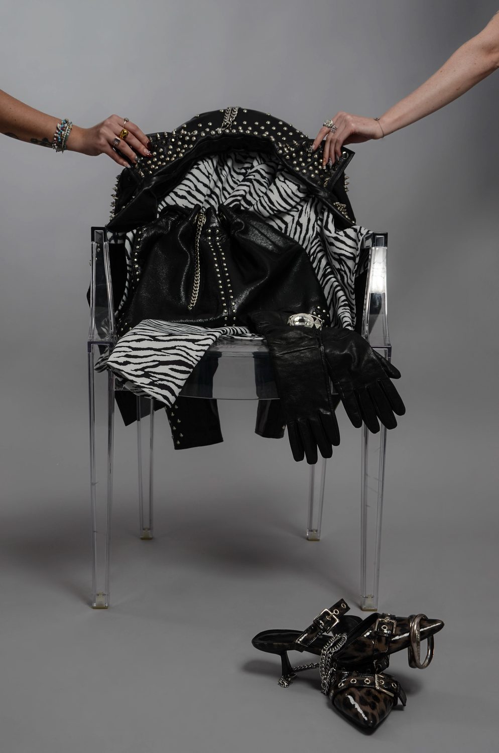 Intervention — two hands enter the frame from above, lifting and spreading the zebra-print fabric across the heaped jacket on the acrylic chair