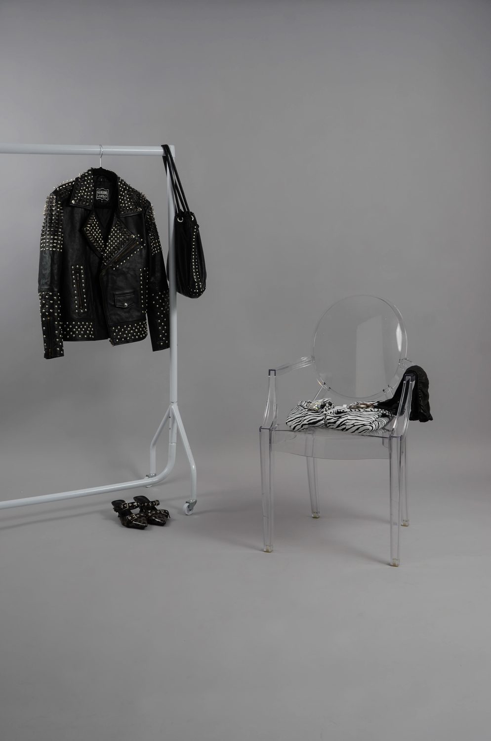 A studded leather jacket and shoulder bag hung on a bare garment rail beside a clear acrylic chair