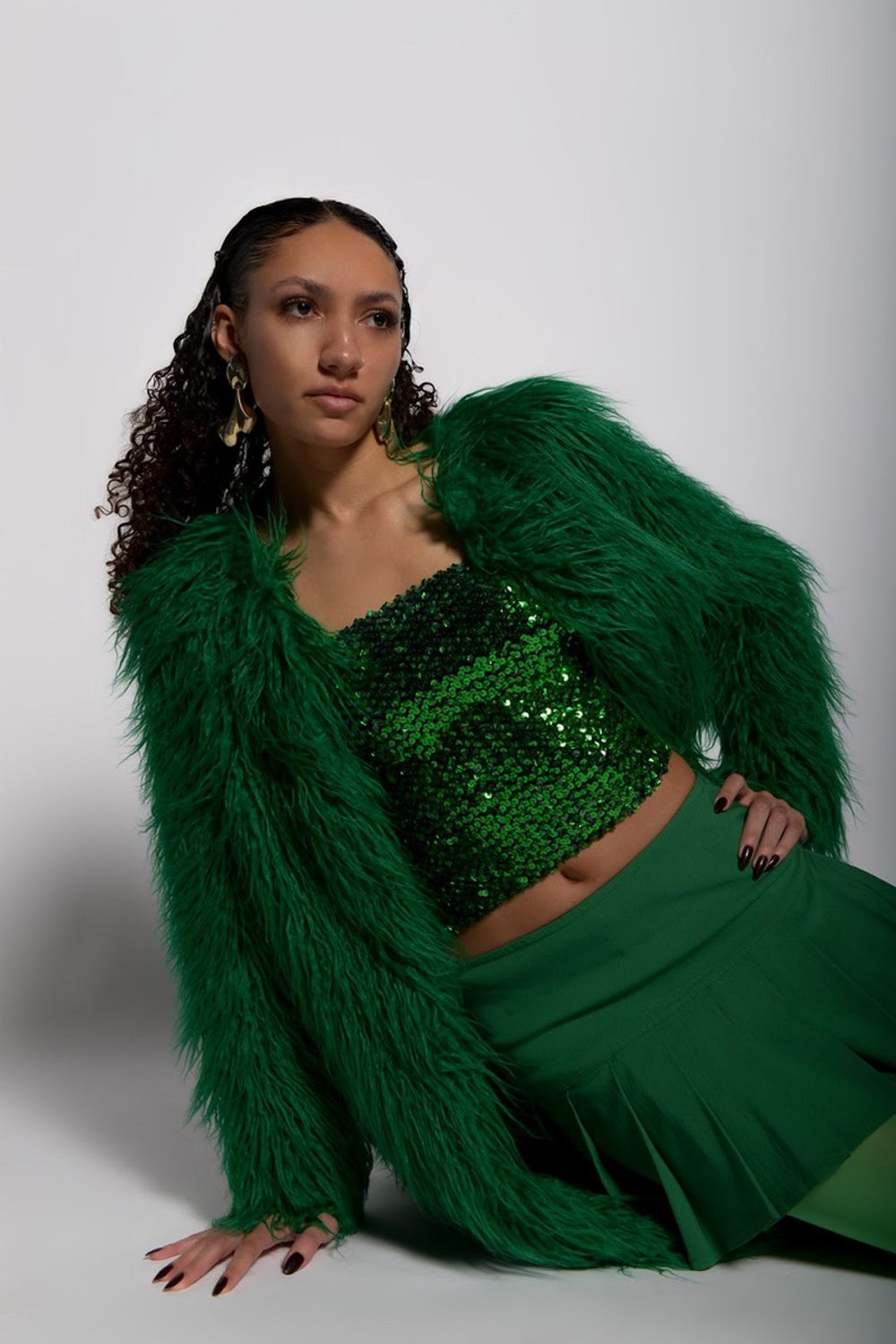 Seated in an emerald shaggy fur coat over green sequins, shot against a pale studio ground