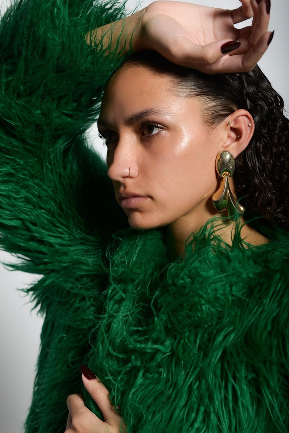 Close portrait framed by emerald fur, one hand raised behind the head, gold statement earring and slicked hair
