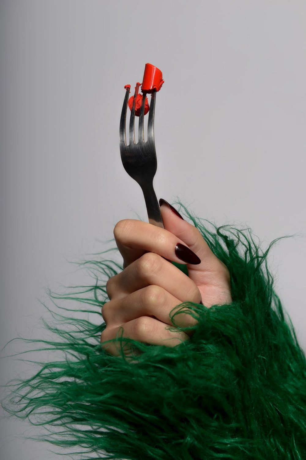 Still life detail — a hand with dark red nails holding a fork against emerald fur, a smear of red at the tines