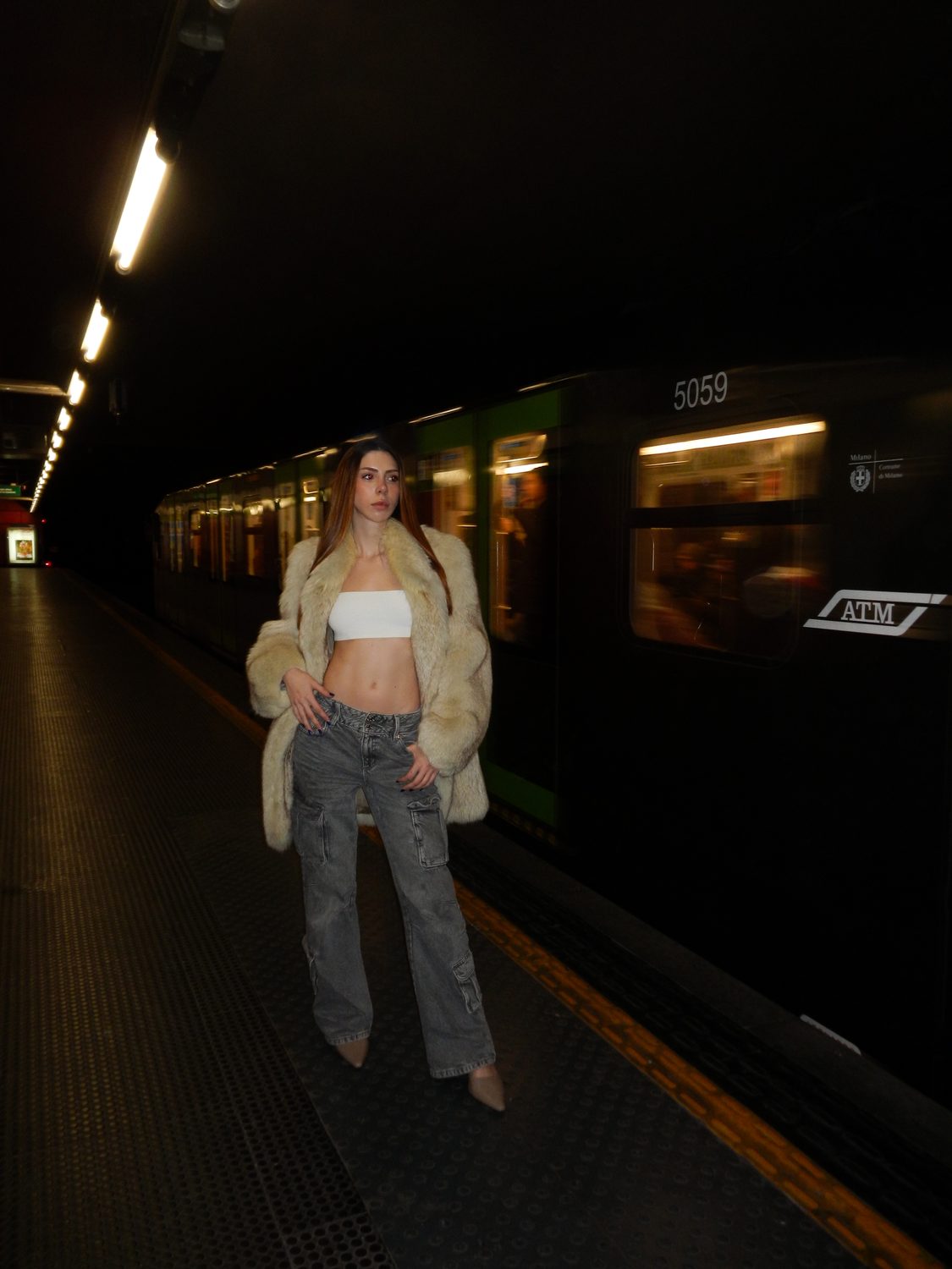 The street-style look on a Milan metro platform at night — a cream shaggy coat over a white top and grey jeans, a train blurred alongside