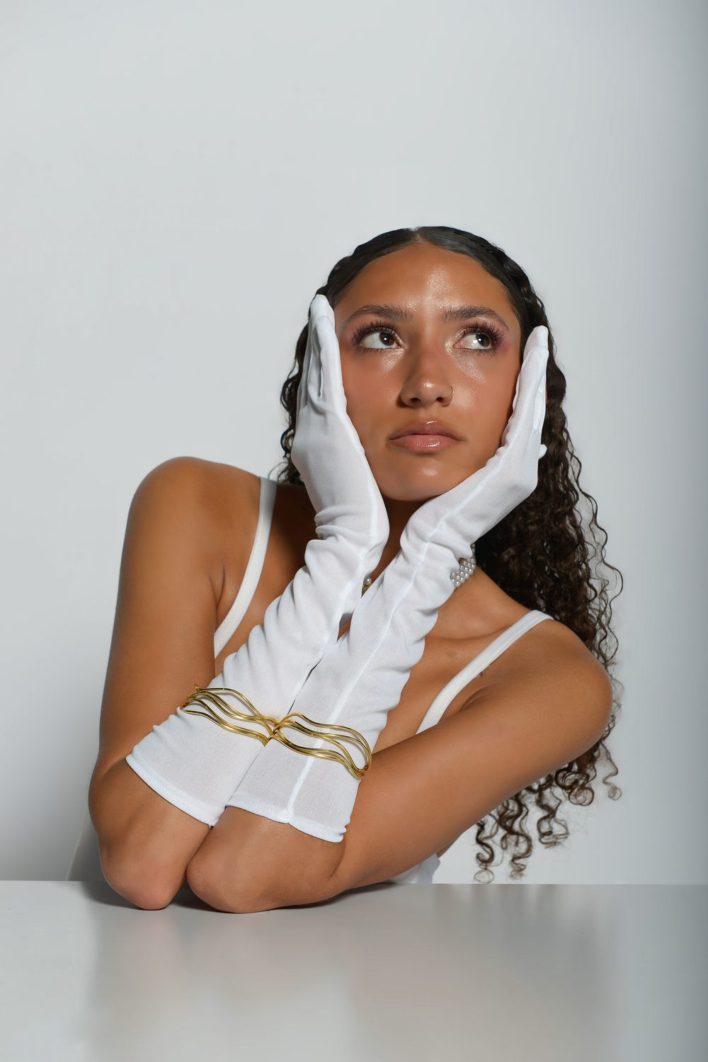 The Harper’s Bazaar styling — seated at a white table, white opera gloves framing the face, pearl choker and gold drop earrings