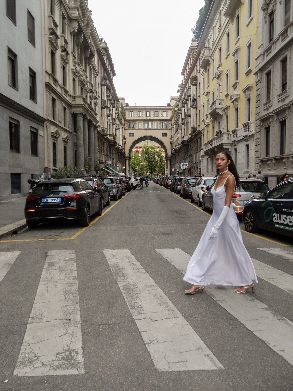 The Harper’s Bazaar styling crossing an empty Milan street at a zebra crossing, parked cars either side and an archway in the distance