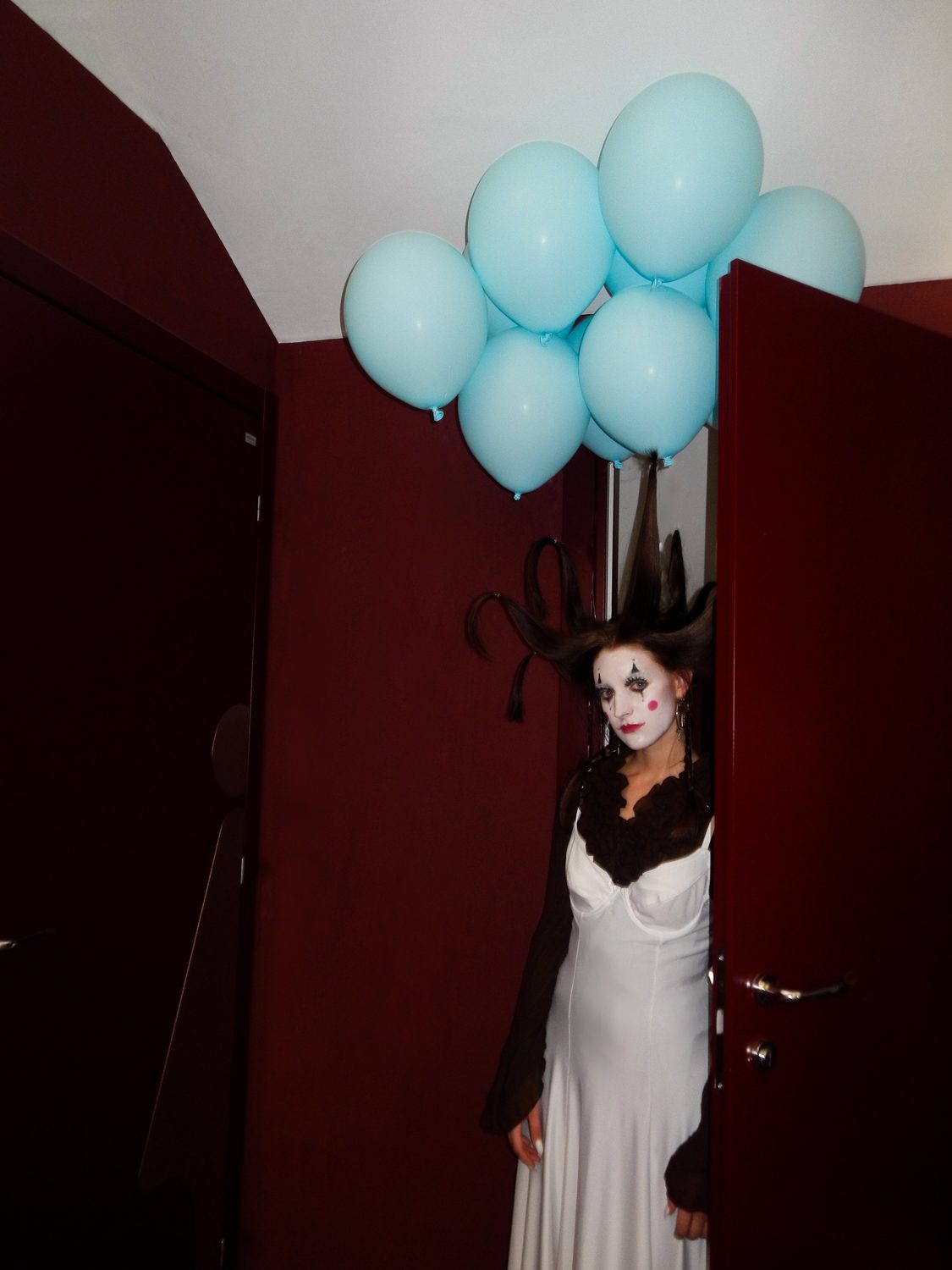 The King Kong styling standing in a deep red doorway holding balloons, the white dress under dark layers