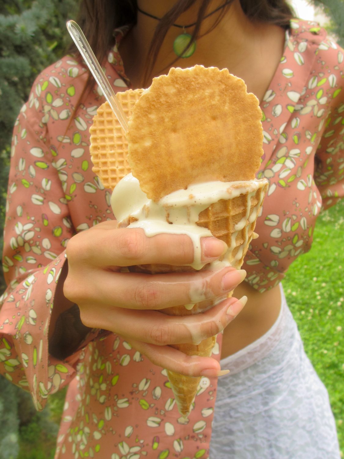 Close outdoor frame — hands holding a pistachio gelato cone up to camera, the pistachio-print shirt and pale skirt behind, grass in the background
