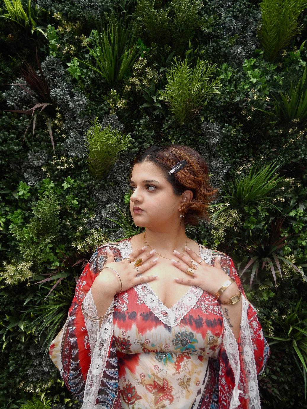 Participant 001 among dense green foliage, hands drawn to the chest, sheer floral print top with lace trim and layered gold jewellery