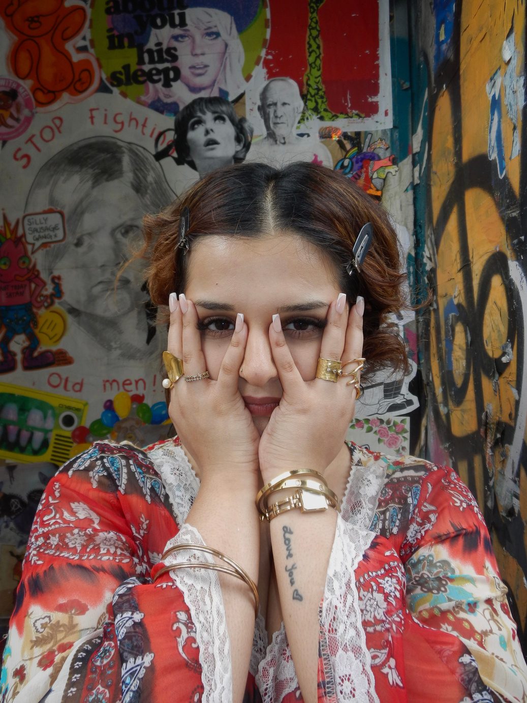 Participant 001 in close portrait, sunglasses on and both hands raised to the face, sheer red and white printed top with stacked gold rings and bangles