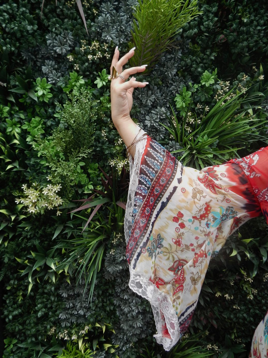 Participant 001 with one arm raised into dense green foliage, the wide sheer printed sleeve falling back from the wrist