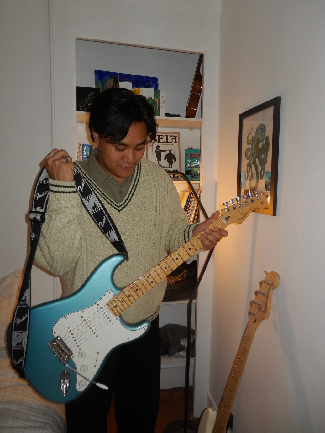 Participant 002 holding a pale blue electric guitar in a lamplit room, cream jumper and dark trousers — the music reference from his archive