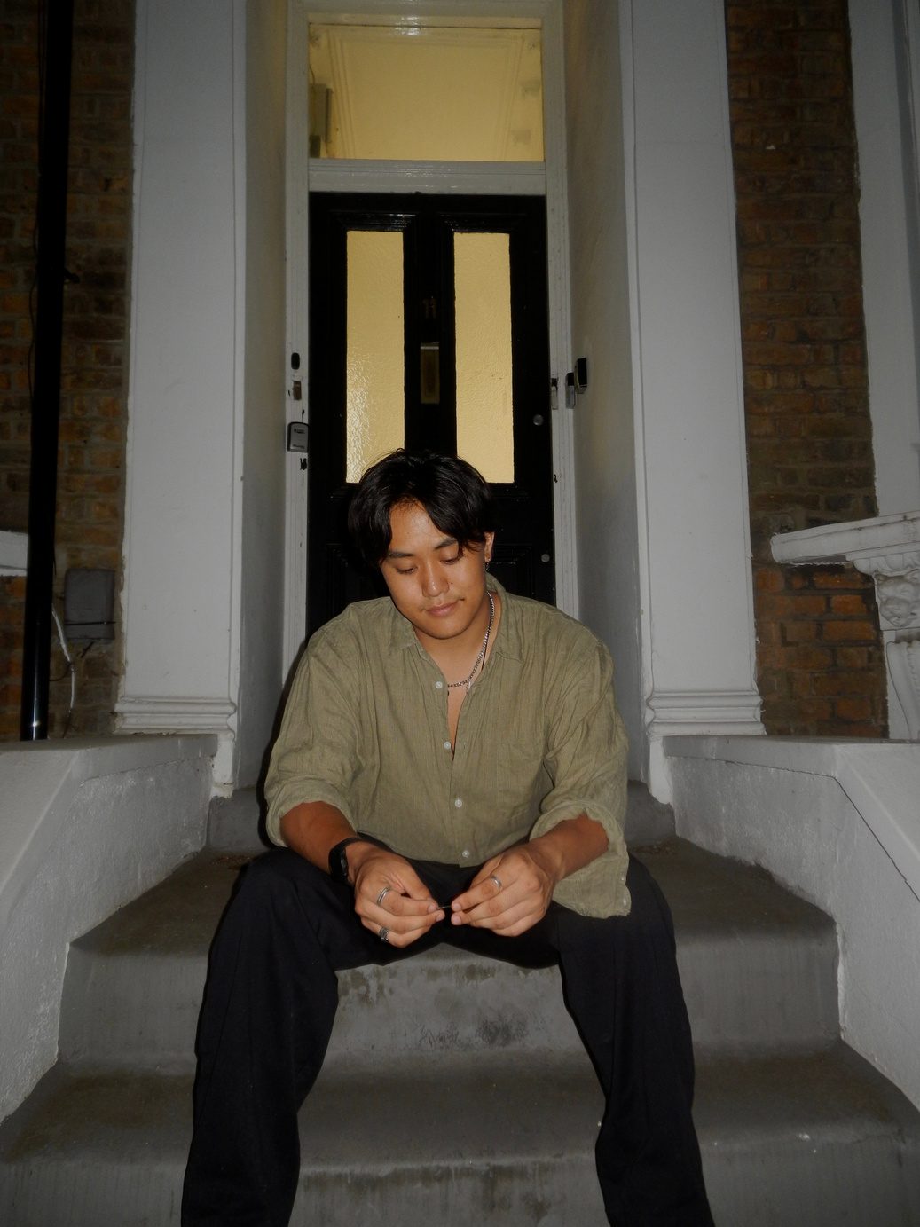 Participant 002 seated in a doorway on stone steps, cream V-neck jumper over a collared shirt, black trousers, hands clasped between the knees