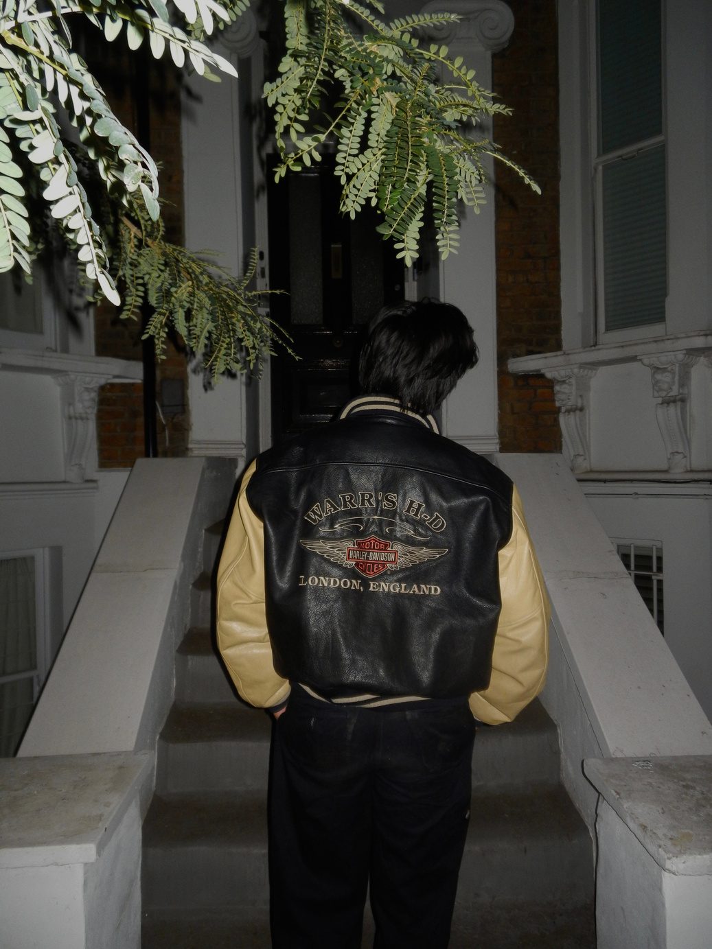 Participant 002 from behind on stone steps beside green foliage, wearing a black and cream varsity jacket