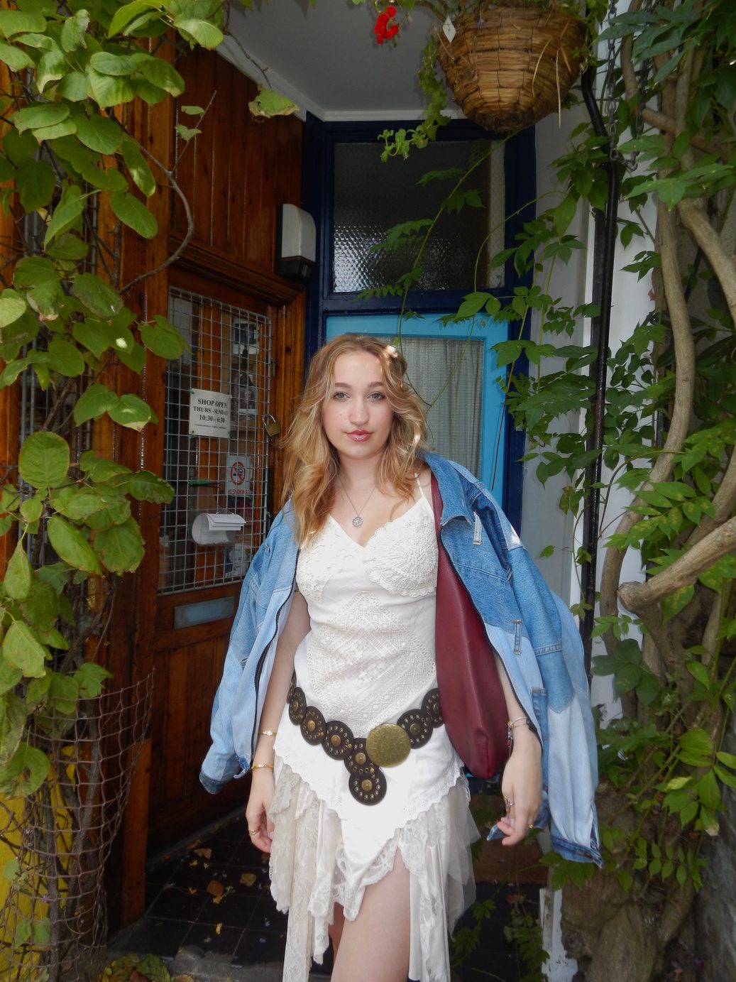 Participant 003 standing in a blue painted doorway framed by ivy, white lace dress with a concho belt and a draped denim jacket