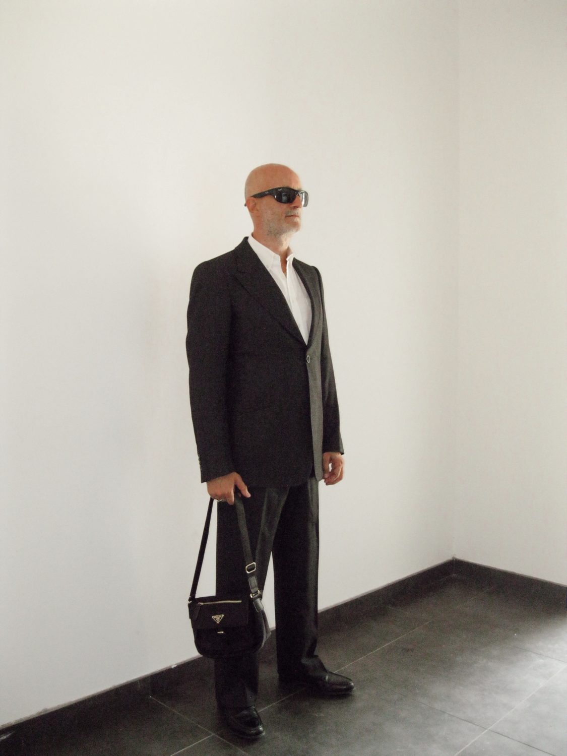 Full length in a bare concrete-floored room — an older male model in charcoal tailoring over a pale knit, dark sunglasses, a black crossbody bag held at the side