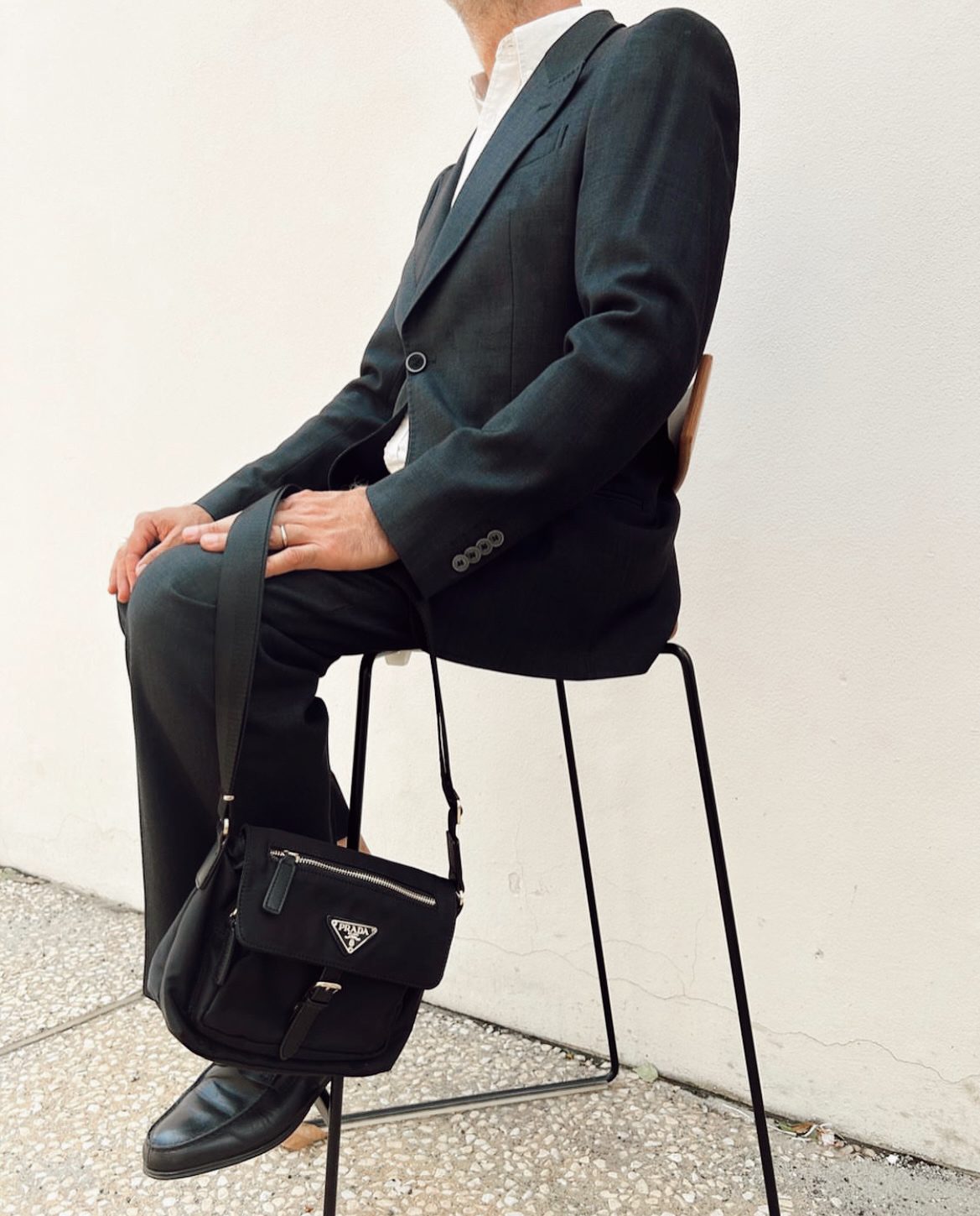 Seated on a high stool against a pale wall, legs crossed, the black crossbody bag hanging low at the side