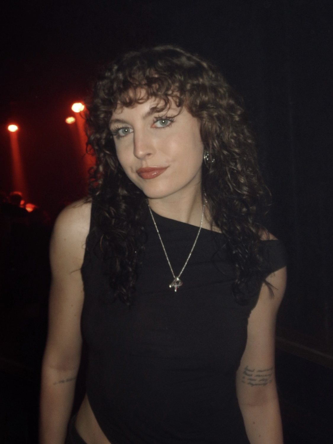 Rachel Riemersma photographed in low light, wearing a black asymmetric top and a fine silver chain, with red stage lights behind her