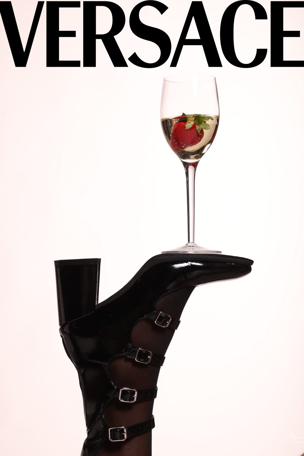 Still life — a black buckled leather boot laid on its side balancing a glass of white wine with a strawberry, against a pale ground