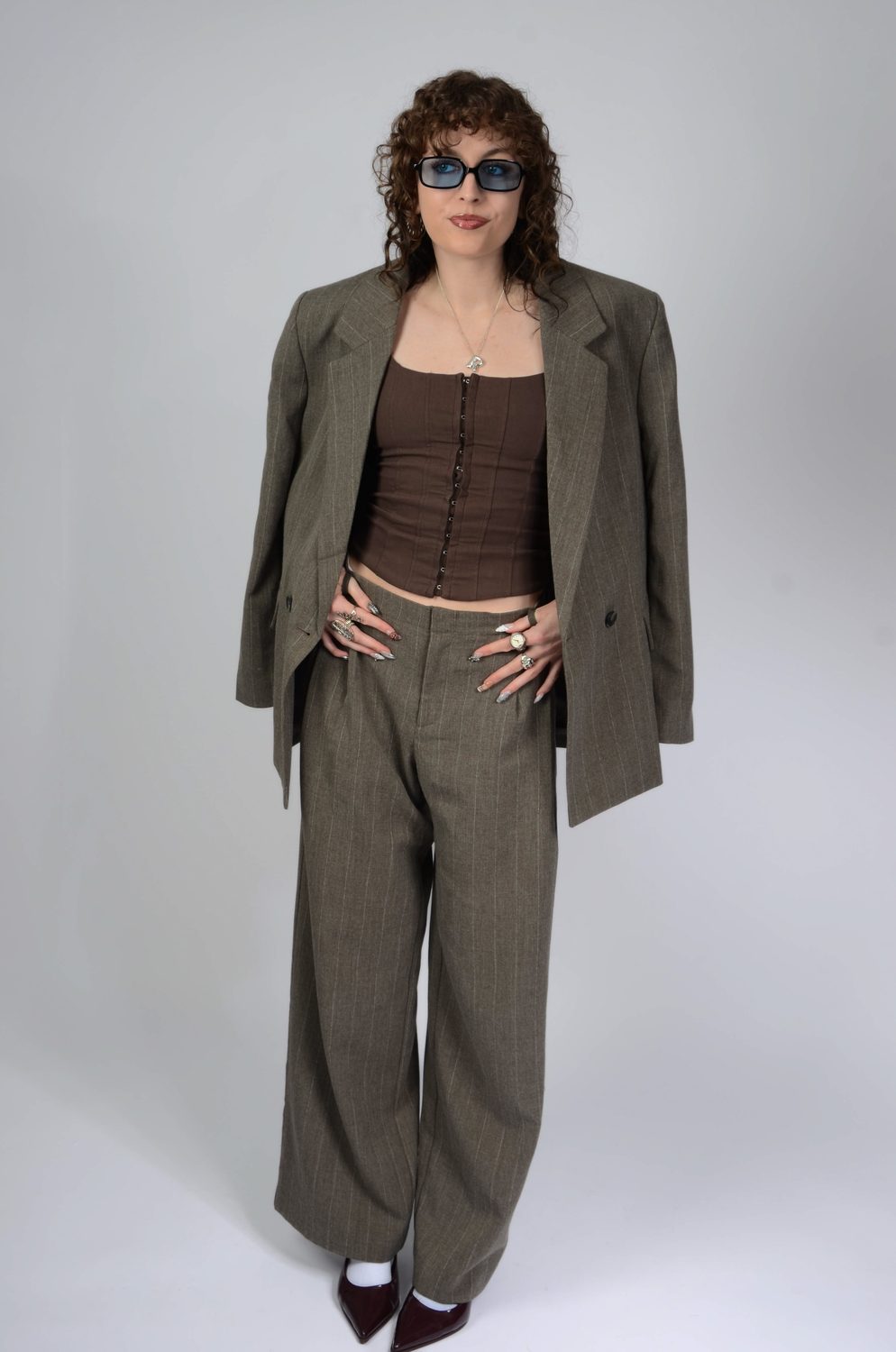 An oversized olive suit worn open over a corset top with blue-tinted glasses, against a pale studio backdrop