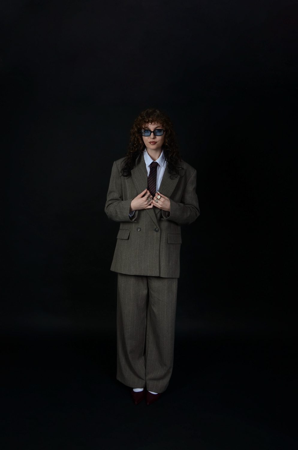 The masculine reading — the suit buttoned over a pale shirt and tie against a black backdrop, blue-tinted glasses on, hands at the jacket front