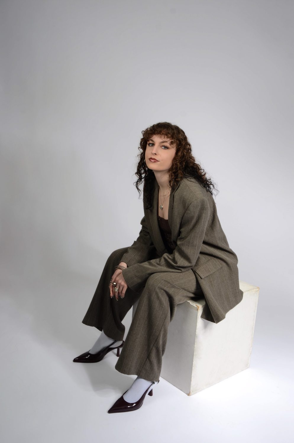The feminine reading seated on a white plinth, legs crossed, the oversized blazer falling open over the corset top, pointed black heels