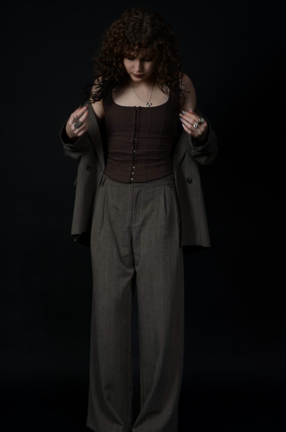 Standing on a black backdrop holding the blazer open at both lapels over the corset top